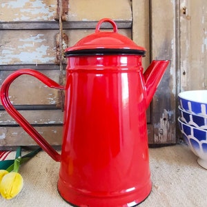 Peut inclure: Une cafetière en émail rouge vif avec un bord et une poignée noirs. La cafetière de style vintage a un couvercle et un bec verseur. La poignée est incurvée, et la cafetière est posée sur un fond en bois rustique.