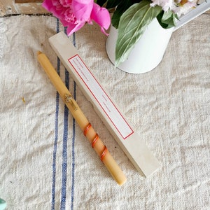 May include: A long, cream-colored candle with a gold cross and red spiral accents, next to a rectangular box with red trim and text. A bouquet of pink peonies and white flowers in a white pitcher are in the background.