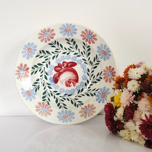 Peut inclure: Une assiette en céramique blanche avec un motif floral. L'assiette présente un motif central rouge et bleu entouré d'une couronne de feuilles vertes et d'une bordure de fleurs bleues et roses alternées. Un bouquet de fleurs colorées est à droite.