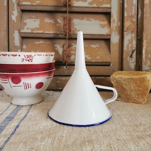 May include: A white enamel funnel with a blue rim and handle rests on a striped cloth. Three red and white patterned bowls are nearby. A block of yellow soap is in the background. The items are in front of a weathered wooden shutter.