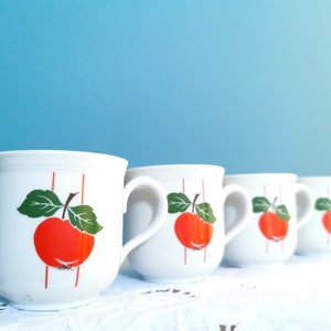Peut inclure: Quatre tasses en céramique blanche avec un motif de pomme rouge et des feuilles vertes. Chaque tasse a une anse et des rayures verticales rouges. Les tasses sont disposées en rangée sur une nappe en dentelle blanche.