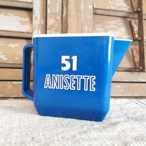 Peut inclure: Pichet en plastique bleu avec un bord, une poignée et un bec verseur blancs. Le pichet porte le numéro "51" et le mot "ANISETTE" en lettres blanches. Il est posé sur un tissu à rayures blanches et bleues, sur un fond en bois vieilli.