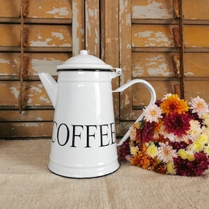 Könnte beinhalten: Eine weiße Emaille-Kaffeekanne mit schwarzem Rand und Griff, das Wort "COFFEE" in schwarzer Schrift. Ein Strauß bunter Blumen liegt neben der Kanne. Der Hintergrund zeigt verwitterte Holzläden.