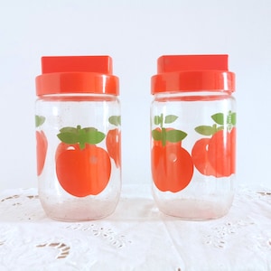 May include: Two clear glass jars with red lids. Each jar is decorated with a repeating pattern of red apples with green leaves. The jars are displayed on a white, lace-covered surface.