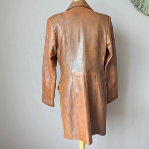 May include: A tan leather coat displayed on a wooden stand. The coat has a classic design with a notched collar, long sleeves, and a tailored fit. The leather appears smooth and has a warm, brown hue. The coat is a mid-length style, suitable for various occasions.