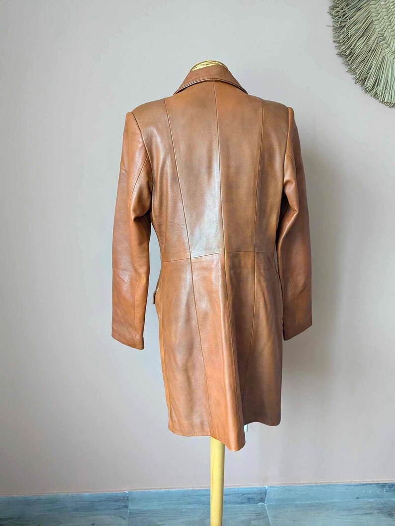 May include: A tan leather coat displayed on a wooden stand. The coat has a classic design with a notched collar, long sleeves, and a tailored fit. The leather appears smooth and has a warm, brown hue. The coat is a mid-length style, suitable for various occasions.