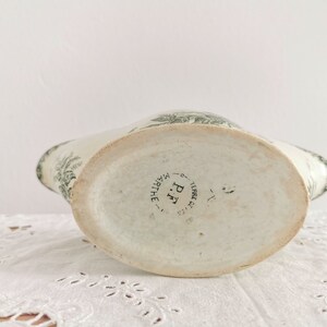 May include: A vintage white ceramic pitcher with a green floral pattern. The pitcher features a wide, oval base and a curved spout. The base of the pitcher is stamped with "P.F. MARTHE TERRE DE FER".