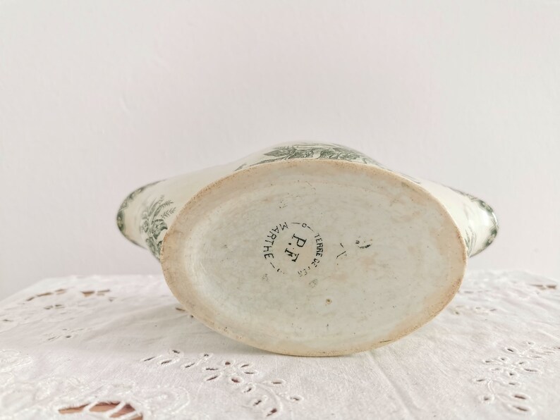 May include: A vintage white ceramic pitcher with a green floral pattern. The pitcher features a wide, oval base and a curved spout. The base of the pitcher is stamped with "P.F. MARTHE TERRE DE FER".