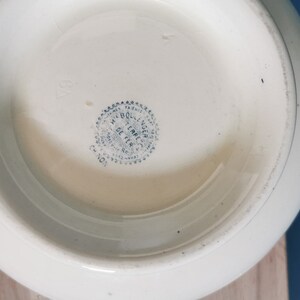 May include: Off-white ceramic bowl with a rounded shape. The bottom of the bowl features a blue and white circular stamp with text. The bowl has a smooth, glossy finish. The bowl is likely vintage or antique.