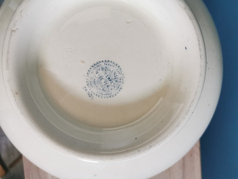 May include: Off-white ceramic bowl with a rounded shape. The bottom of the bowl features a blue and white circular stamp with text. The bowl has a smooth, glossy finish. The bowl is likely vintage or antique.