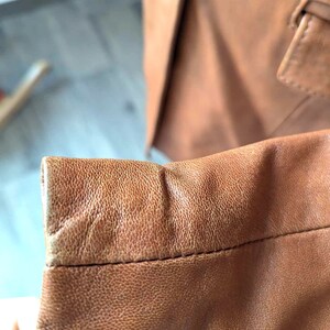May include: Close-up of a brown leather blazer sleeve, showcasing the textured surface and stitching details. The sleeve is a warm, medium brown color, with a slightly worn appearance. The blazer's button and lapel are visible in the background.