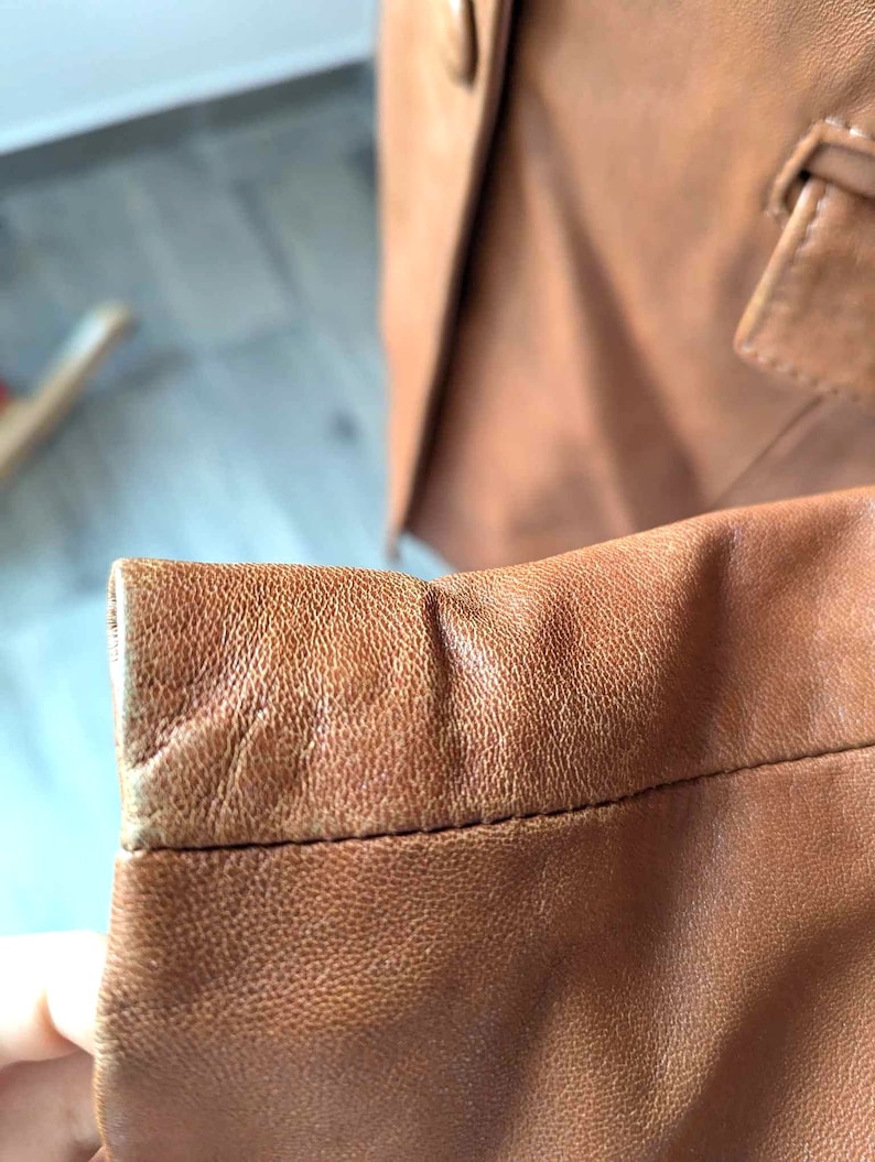 May include: Close-up of a brown leather blazer sleeve, showcasing the textured surface and stitching details. The sleeve is a warm, medium brown color, with a slightly worn appearance. The blazer's button and lapel are visible in the background.