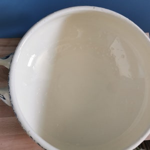 May include: Cream-colored ceramic serving bowl with two handles and a blue floral design around the rim. The bowl has a wide, open shape and a glossy finish, suitable for serving soups or stews.