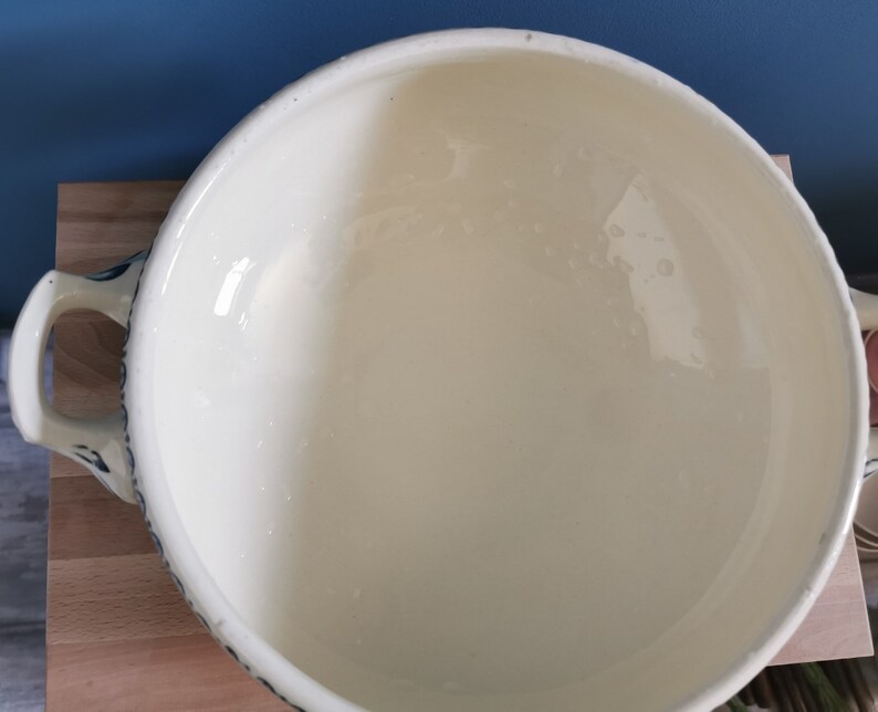 May include: Cream-colored ceramic serving bowl with two handles and a blue floral design around the rim. The bowl has a wide, open shape and a glossy finish, suitable for serving soups or stews.