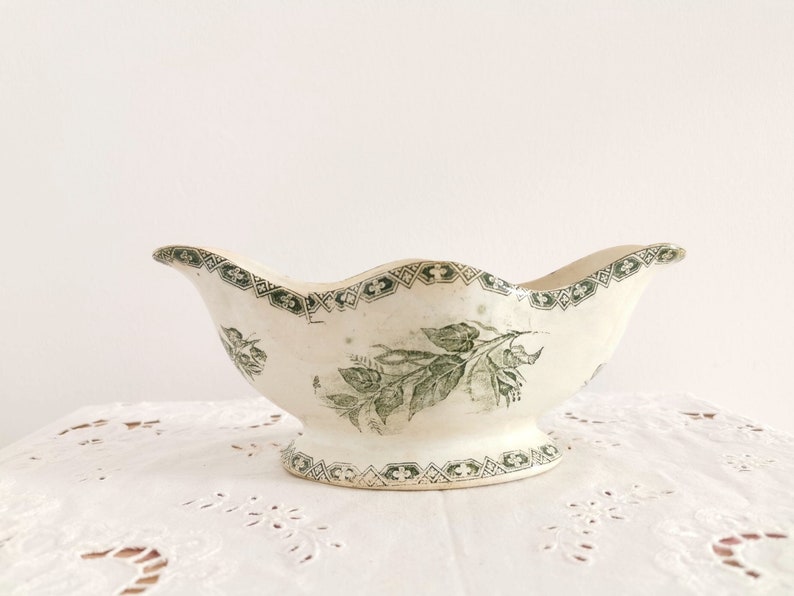 May include: Vintage cream-coloured gravy boat with a scalloped rim and pedestal base. It features a green floral and leaf pattern, with a decorative green border. The design is reminiscent of antique tableware, adding a touch of elegance to any table setting.
