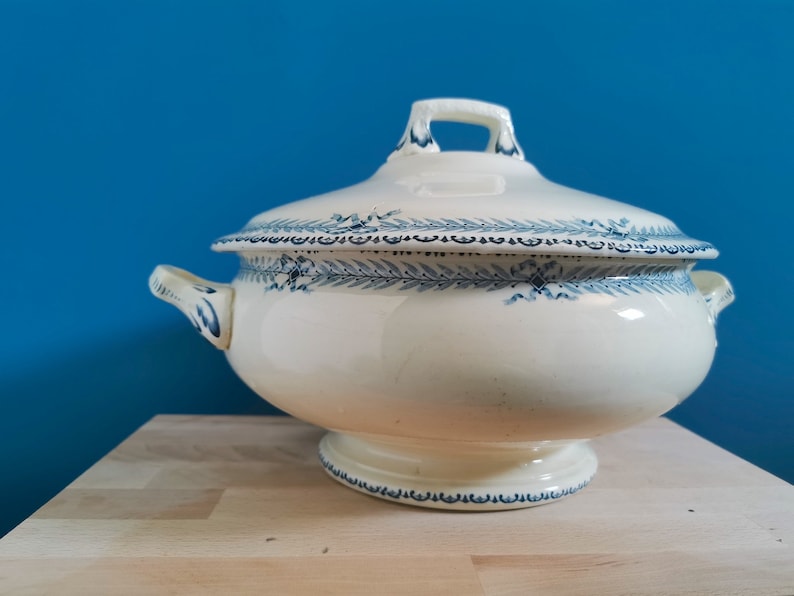 May include: A white ceramic soup tureen with a lid, featuring blue floral and leaf patterns. The tureen has two side handles and a handle on the lid. It is set on a pedestal base, perfect for serving soups and stews.