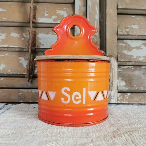 May include: Vintage orange enamelware salt container with a decorative top. The container features the word "Sel" in white lettering, with a geometric design. The container is cylindrical and has a wooden lid.