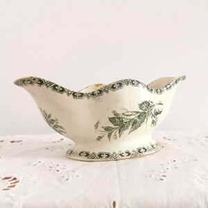 May include: Cream ceramic gravy boat with a scalloped rim and pedestal base. It features a green floral and geometric pattern. The design includes a detailed botanical illustration and decorative border, adding a vintage aesthetic.