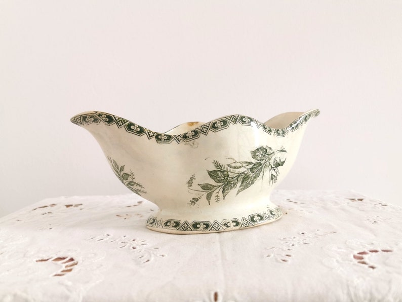 May include: Cream ceramic gravy boat with a scalloped rim and pedestal base. It features a green floral and geometric pattern. The design includes a detailed botanical illustration and decorative border, adding a vintage aesthetic.