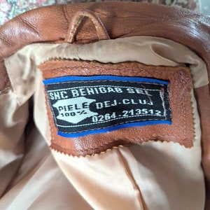May include: A brown leather jacket with a light tan lining. The jacket has a small loop at the collar and a tag with text in black and blue. The tag reads "PIELE DEJ-CLUJ 100%" and a phone number. The jacket appears to be vintage.