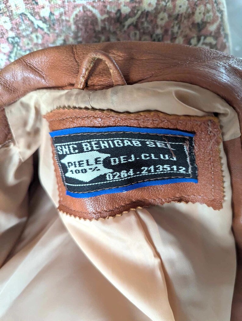 May include: A brown leather jacket with a light tan lining. The jacket has a small loop at the collar and a tag with text in black and blue. The tag reads "PIELE DEJ-CLUJ 100%" and a phone number. The jacket appears to be vintage.