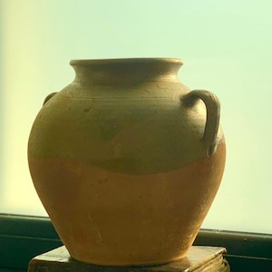 Peut inclure: Un pot en terre cuite rustique à deux tons avec deux poignées incurvées. Le pot est de couleur marron chaud en bas, passant à un brun verdâtre en haut. Il repose sur un livre sombre et vieilli.