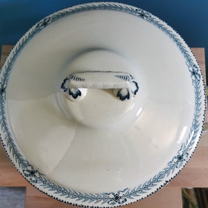 May include: Cream-colored, lidded serving dish with a blue floral border and handles. The lid features a central handle and a decorative blue design. The dish is set on a wooden surface, suggesting a kitchen or dining setting.