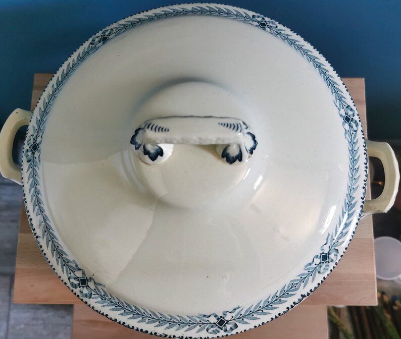 May include: Cream-colored, lidded serving dish with a blue floral border and handles. The lid features a central handle and a decorative blue design. The dish is set on a wooden surface, suggesting a kitchen or dining setting.