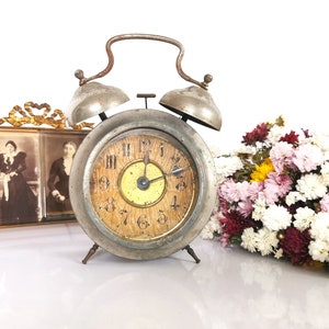 Peut inclure: Un réveil argenté ancien avec un cadran en bois vieilli et des chiffres romains. La montre a deux cloches sur le dessus et repose sur trois petits pieds. Une photographie encadrée et un bouquet de fleurs sont en arrière-plan.