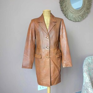 May include: A brown leather blazer with a classic design. The blazer features a notched collar, two front pockets, and three gold-colored buttons. The jacket is displayed on a wooden mannequin.