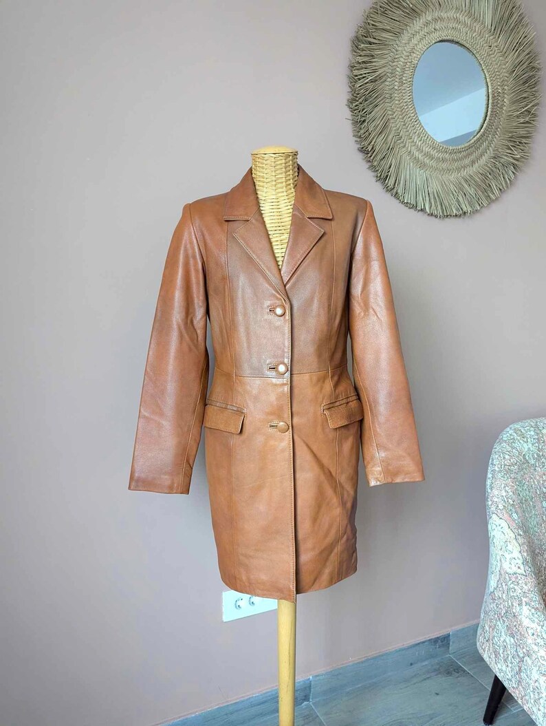 May include: A brown leather blazer with a classic design. The blazer features a notched collar, two front pockets, and three gold-colored buttons. The jacket is displayed on a wooden mannequin.