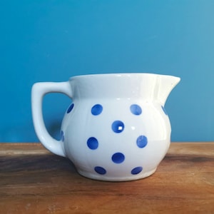May include: A white ceramic jug with a handle and spout, decorated with blue polka dots. The jug is positioned on a wooden surface against a blue backdrop. This decorative jug is suitable for serving beverages.