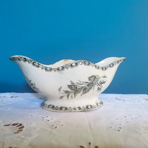 May include: A white porcelain gravy boat with a scalloped rim and pedestal base. It has a black and white geometric border and floral design. The gravy boat is on a white lace tablecloth against a blue background.