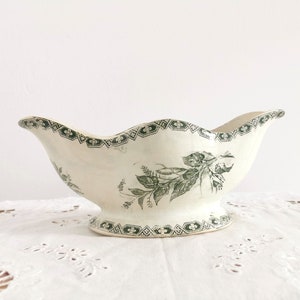May include: Vintage cream-coloured gravy boat with a scalloped rim and pedestal base. It has a green floral pattern and geometric border. A classic piece for serving sauces or dressings.