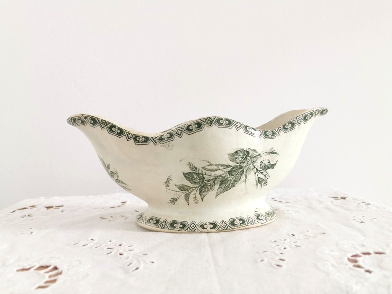May include: Vintage cream-coloured gravy boat with a scalloped rim and pedestal base. It has a green floral pattern and geometric border. A classic piece for serving sauces or dressings.