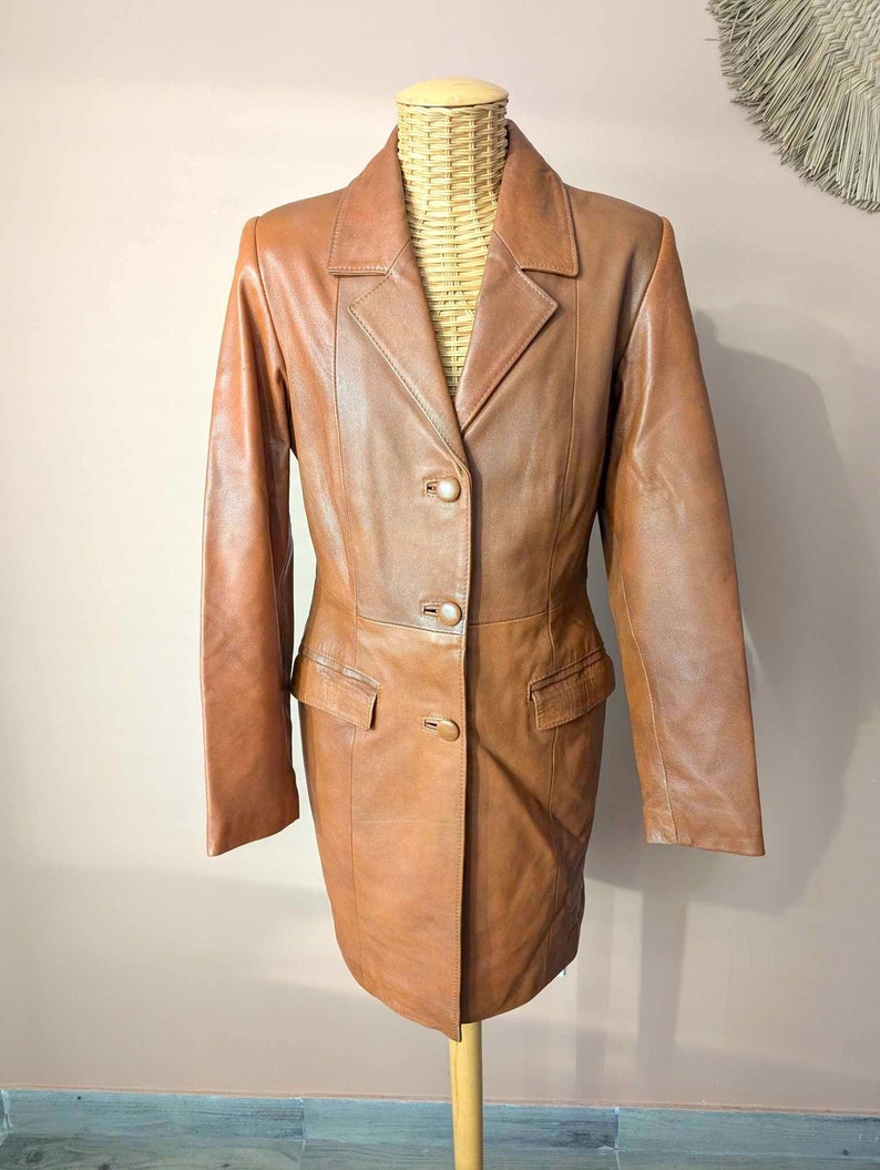 May include: A brown leather blazer with a classic design. The blazer features a notched collar, two front pockets, and three button closure. The jacket is displayed on a mannequin.