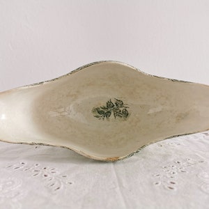 May include: An antique, oblong serving dish with a cream interior and a green floral design. The rim has a delicate green pattern. The dish is on a white, textured surface, suggesting a vintage aesthetic. The dish is likely made of ceramic.