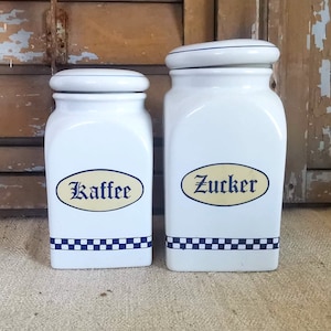 Peut inclure: Deux pots en céramique blancs avec couvercles. L'un porte l'inscription "Kaffee" en bleu sur un ovale jaune, l'autre "Zucker". Les deux ont une bande à carreaux bleus et blancs à la base, pour la cuisine.