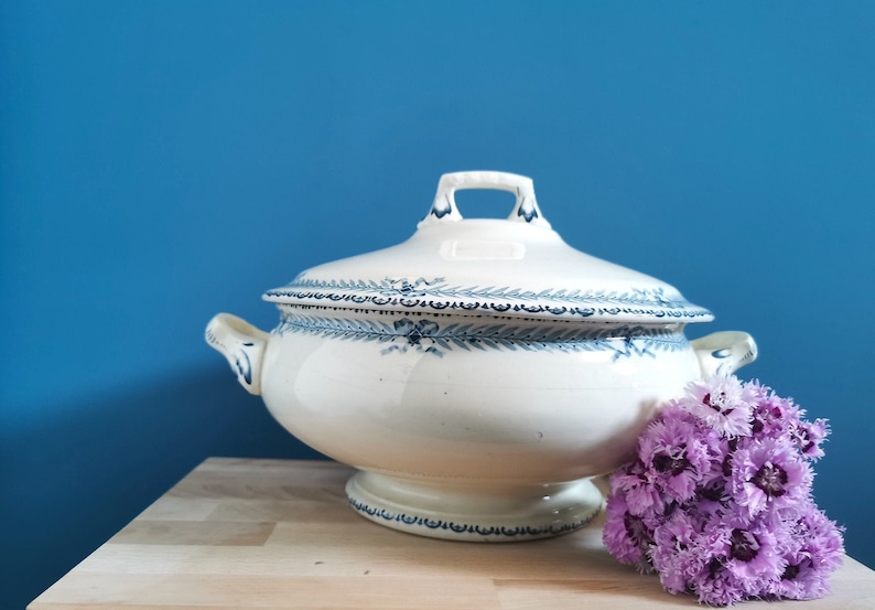 May include: A white ceramic tureen with a lid, adorned with blue floral patterns. The tureen has two handles and a pedestal base. A small bouquet of purple flowers rests beside it on a wooden surface, against a blue backdrop.
