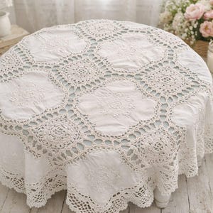 May include: A round white tablecloth with an intricate crochet lace design. The tablecloth is draped over a round table, with a detailed floral pattern in the center and around the edges. The table is surrounded by other decorative items.