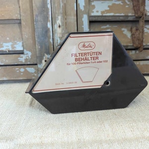 Puede incluir: Un portafiltros de café Melitta negro y marrón. El soporte es trapezoidal con una etiqueta blanca y marrón que dice "FILTERTUTEN BEHÄLTER" y "für 100 Filtertüten 1x4 oder 102". El fondo es una puerta de madera desgastada.