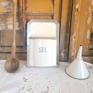 May include: A white enamel salt container with the word "SEL" in blue lettering, a wooden ladle, and a white funnel. The items are arranged on a white lace tablecloth against a rustic wooden background.