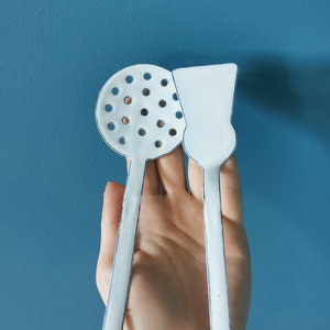 May include: A pair of white kitchen utensils, a slotted spoon and a spatula, are held against a blue backdrop. The utensils have a simple design with black outlines. The slotted spoon has multiple small holes.