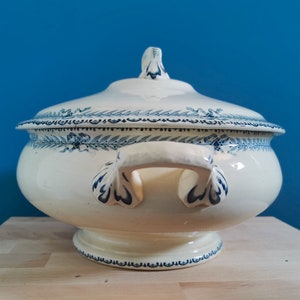 May include: Cream-colored ceramic soup tureen with a lid, adorned with blue floral and leaf patterns. The tureen has two handles and a pedestal base. The lid features a decorative finial. A classic piece for serving soups and stews.