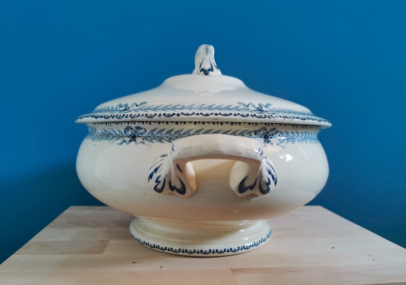 May include: Cream-colored ceramic soup tureen with a lid, adorned with blue floral and leaf patterns. The tureen has two handles and a pedestal base. The lid features a decorative finial. A classic piece for serving soups and stews.