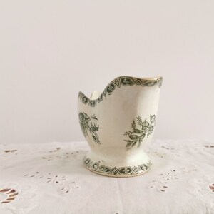 May include: A vintage white ceramic egg cup with a pedestal base. The egg cup has a green floral and geometric pattern around the rim and base. The interior is smooth, and the exterior has a slightly textured surface.