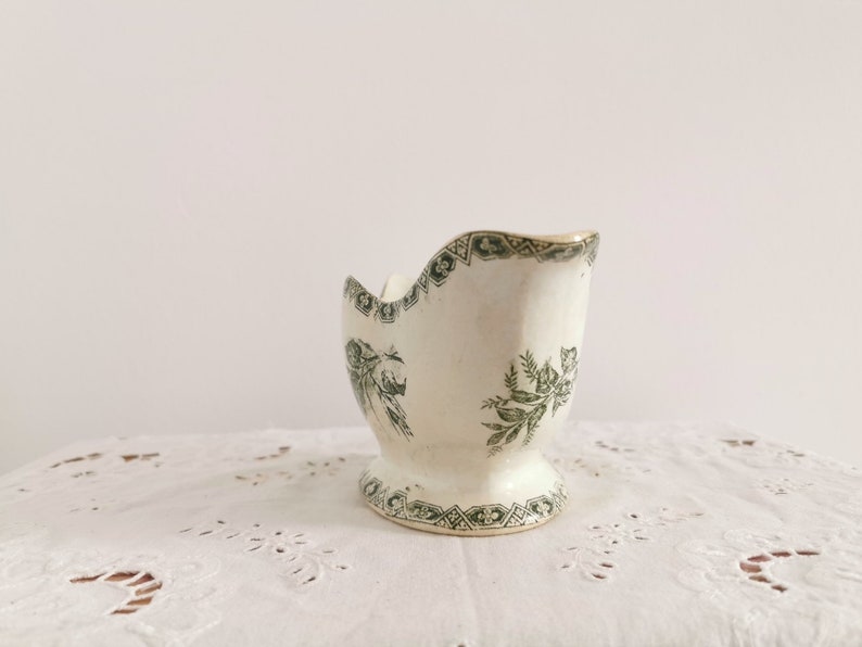 May include: A vintage white ceramic egg cup with a pedestal base. The egg cup has a green floral and geometric pattern around the rim and base. The interior is smooth, and the exterior has a slightly textured surface.