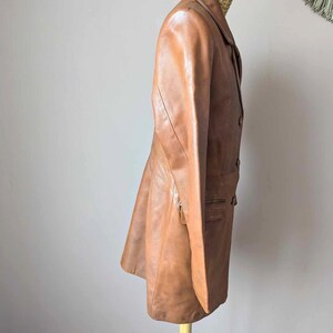 May include: A tan leather blazer with a classic lapel collar and button closure. The blazer features a tailored fit, a front pocket, and a zipper detail. The jacket is displayed on a wooden stand.
