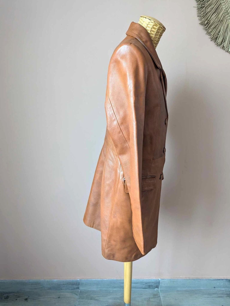 May include: A tan leather blazer with a classic lapel collar and button closure. The blazer features a tailored fit, a front pocket, and a zipper detail. The jacket is displayed on a wooden stand.