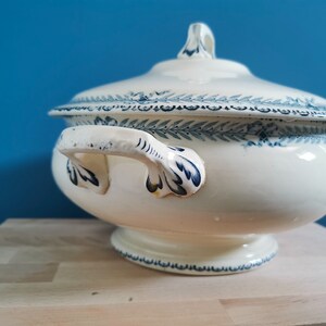May include: A white ceramic tureen with a lid, featuring blue floral and leaf patterns. The tureen has two handles and a pedestal base. The lid has a small handle. The background is a solid blue.